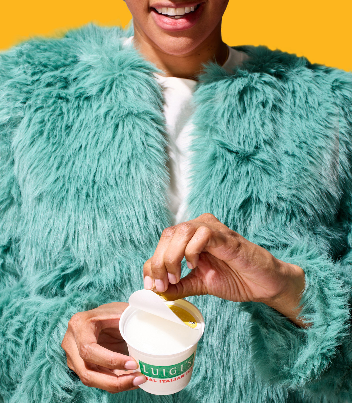 Image of woman opening a cup of lemon LUIGI'S Real Italian Ice. Woman is wearing a blue faux fur jacket. Background is yellow.