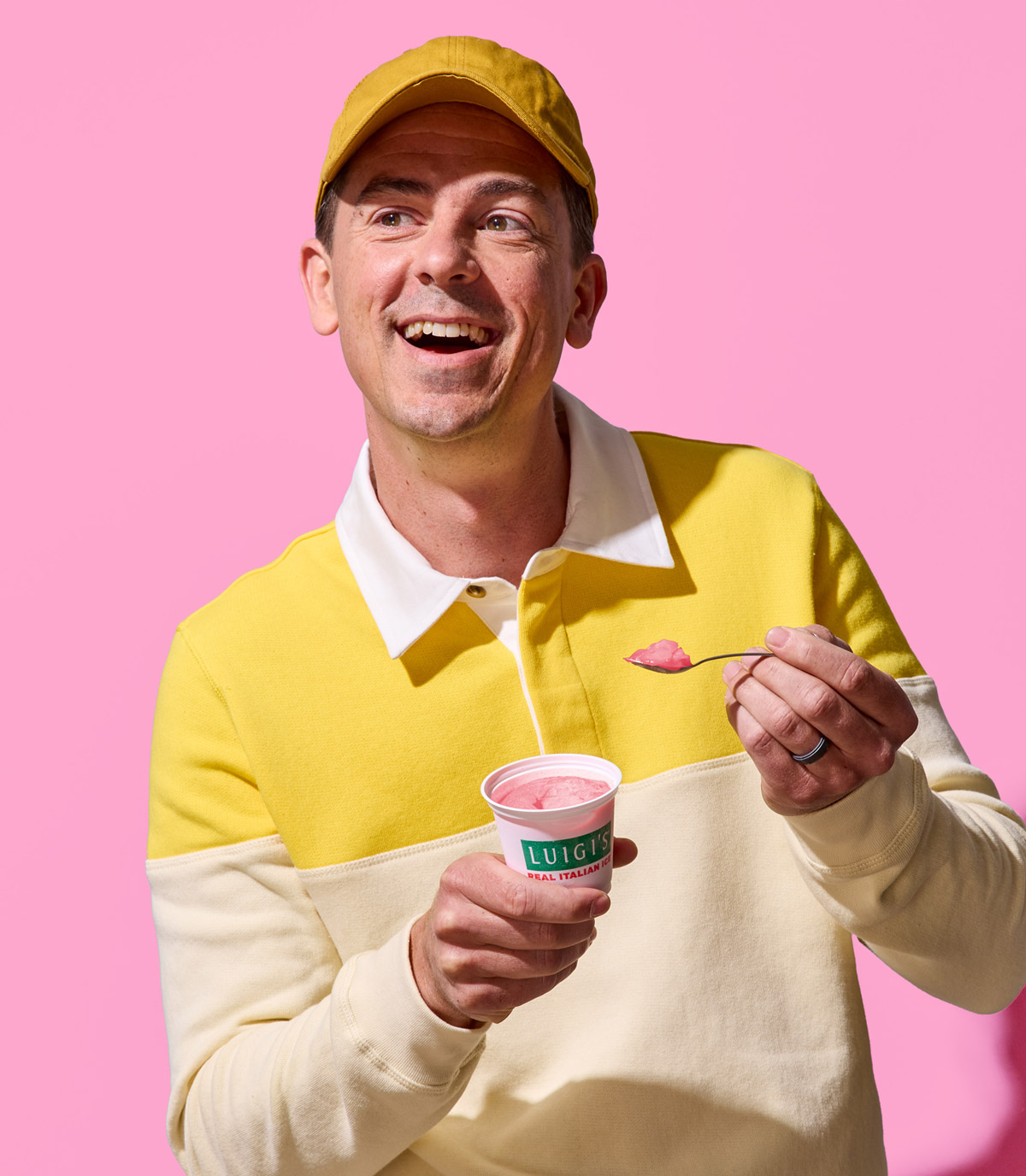 Man wearing yellow shirt and yellow hat eating cherry lemon swirl LUIGI'S Real Italian Ice. Background is pink. Man is smiling and holding cup of Italian ice and spoon.