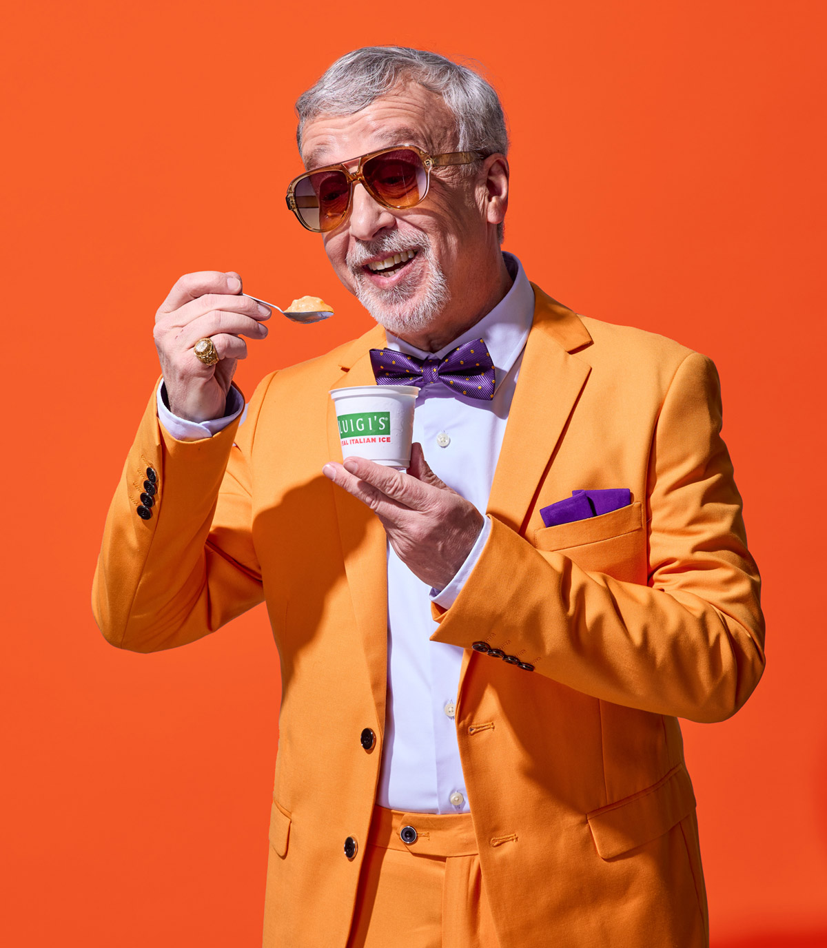 Suave older man, eating mango LUIGI'S Real Italian Ice. Man is in an orange suit with purple bow tie and purple pocket square. He is wearing sunglasses and a pinkie ring. Background is a darker orange.