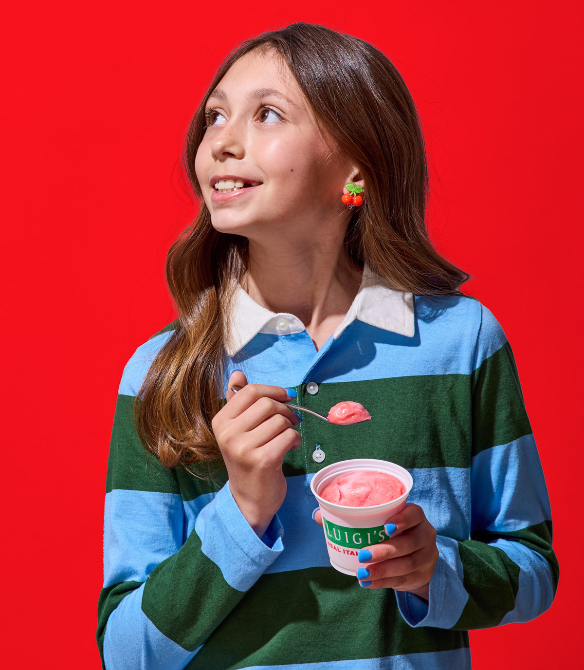 Young girl eating cherry LUIGI'S Real Italian Ice. She is wearing a blue and green rugby shirt and cherry earrings. She's looking over her shoulder, smiling, and holding the cup and spoon in her hand with cherry Italian ice. Background is red.