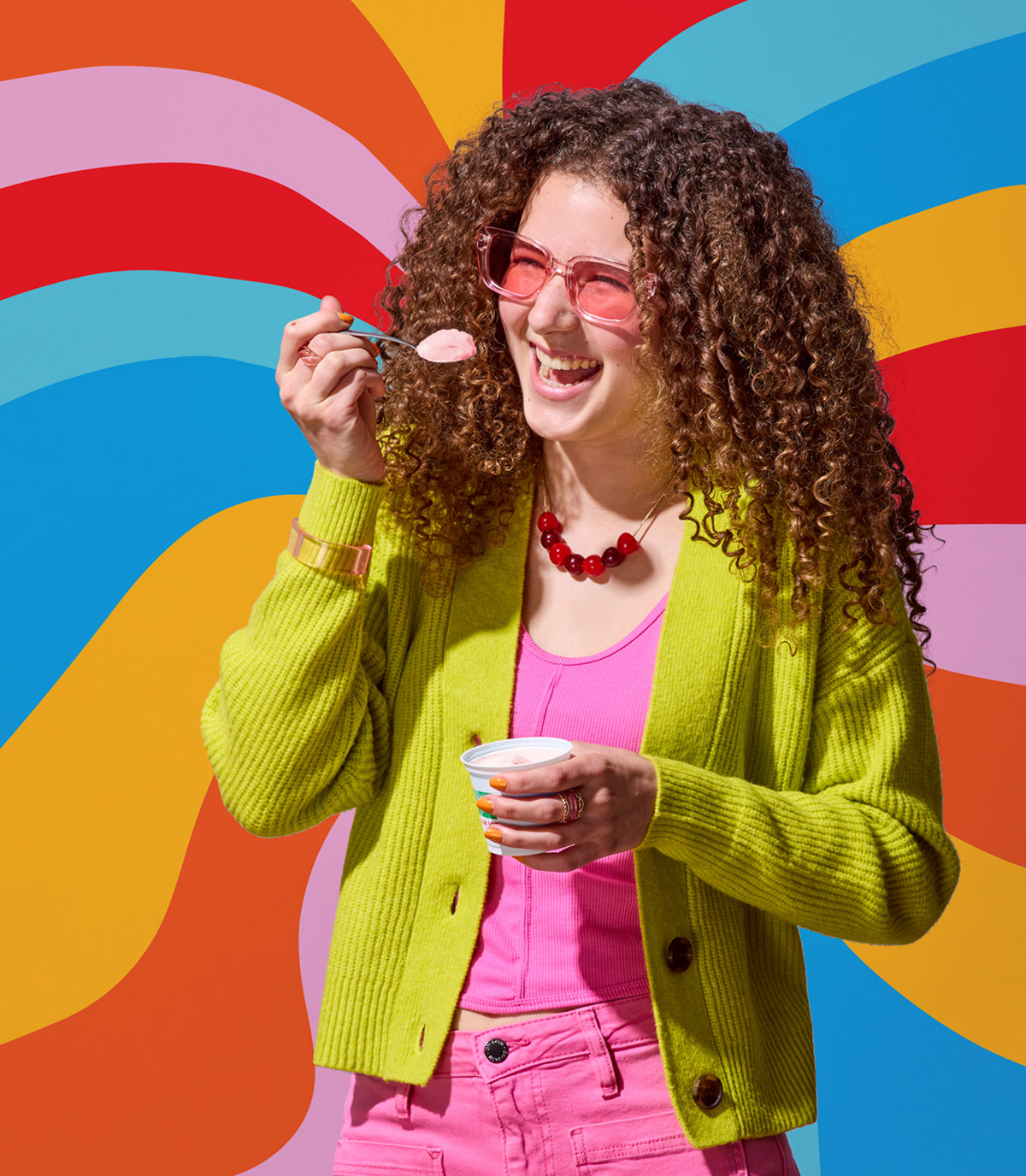 Woman with curly hair is eating kiwi strawberry LUIGI'S Real Italian Ice. Woman is wearing pink sunglasses, a pink outfit, and green sweater. Background is psychedelic rainbow swirls.