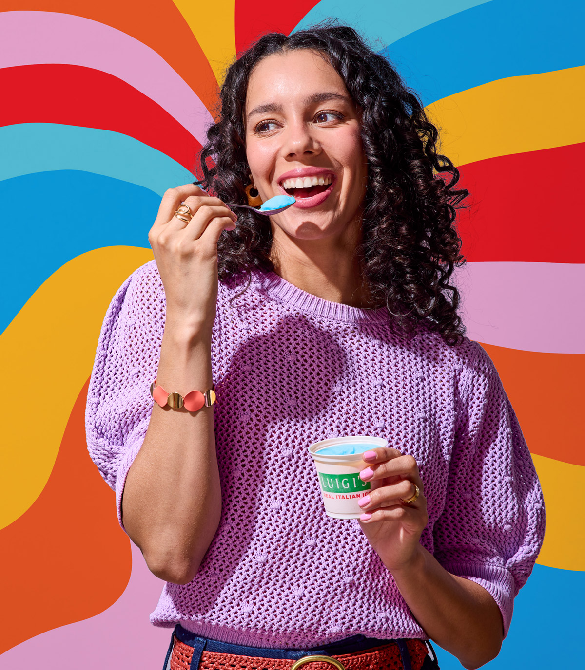 Woman eating blue raspberry LUIGI'S Real Italian Ice. Woman is smiling, wearing a purple top, and is holding the spoon of blue raspberry Italian ice up to her mouth. She is standing in front of a psychedelic rainbow background.