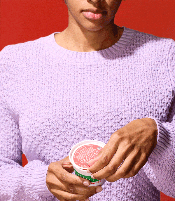 Gif of a woman in purple sweater opening a cup of strawberry LUIGI'S Real Italian Ice. Only the bottom portion of her face is seen, and she is smiling when she opens the cup of Italian ice. The Italian ice has a smile carved in it. Background is red.
