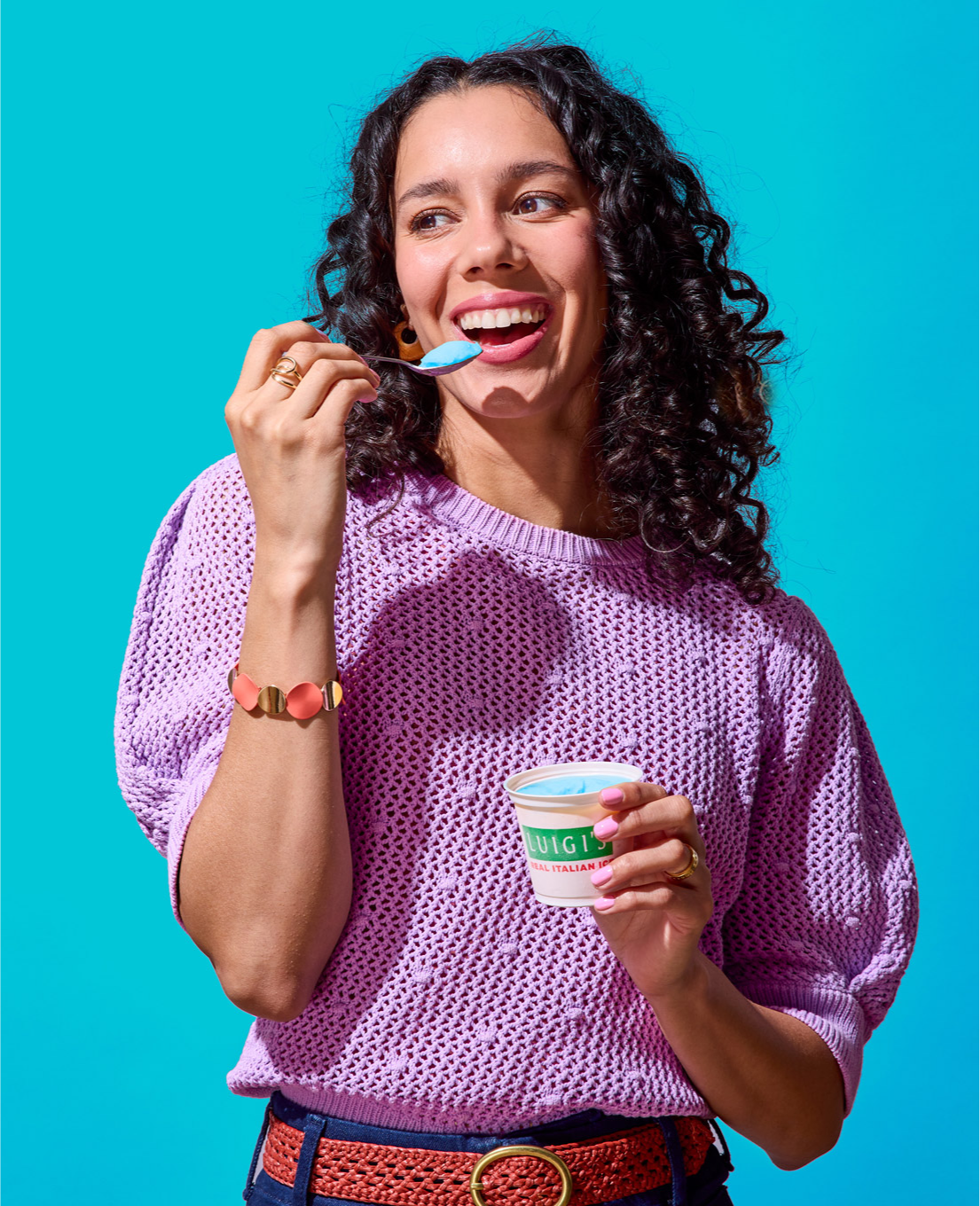 Woman eating blue raspberry LUIGI'S Real Italian Ice. Woman is smiling, wearing a purple top, and is holding the spoon of blue raspberry Italian ice up to her mouth.