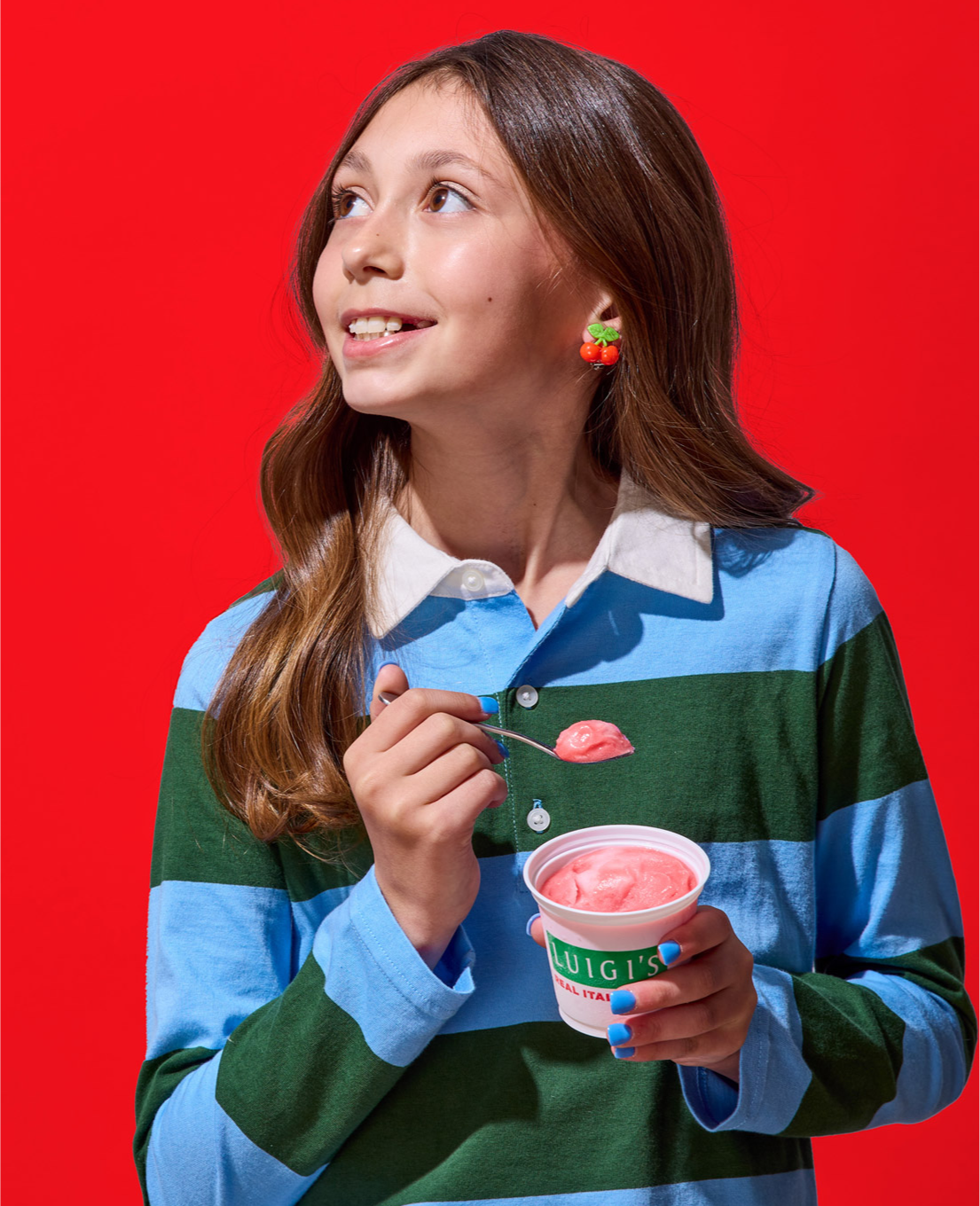 Young girl eating cherry LUIGI'S Real Italian Ice. She is wearing a blue and green rugby shirt and cherry earrings. She's looking over her shoulder, smiling, and holding the cup and spoon in her hand with cherry Italian ice.