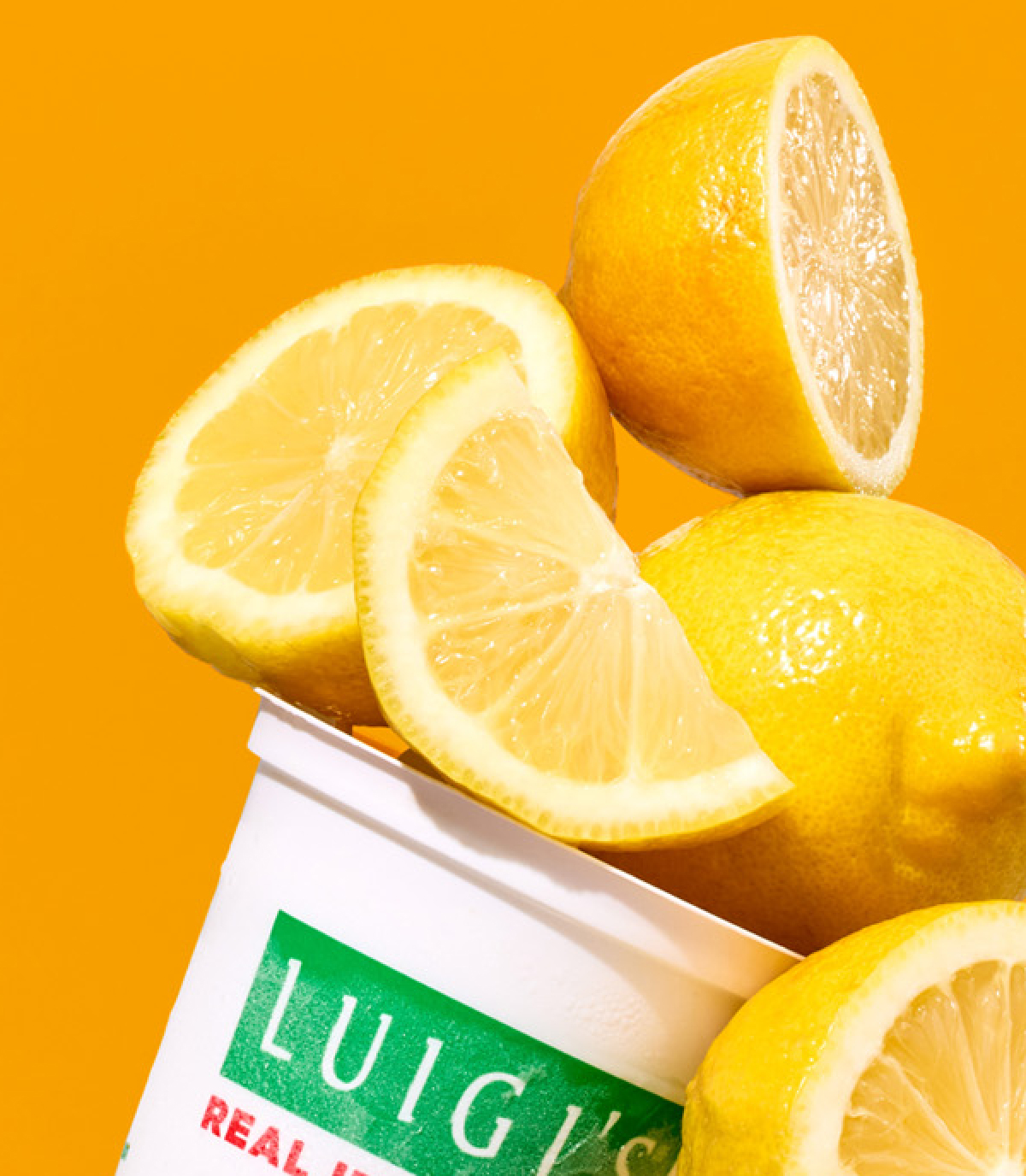 Cropped in close up of a LUIGI'S Real Italian Ice with lemon slices piled on top. Background is yellow.